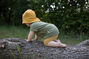 Kleinkind klettert auf einem Baumstamm und hat sommerliche Kleidung aus Naturfasern an